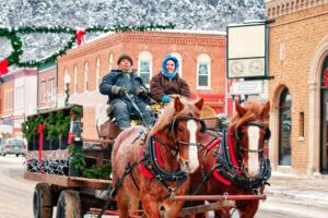 A horse-drawn carriage makes its way down Lanesboro’s snow-covered main street, driven by two warmly dressed locals. The decorated wagon, holiday wreaths, and twinkling lights create a festive small-town winter scene.