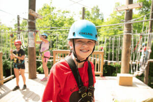 Smiling child wearing a helmet and harness at the Eagle Bluff high ropes course in Lanesboro, with other participants navigating rope bridges in the background.