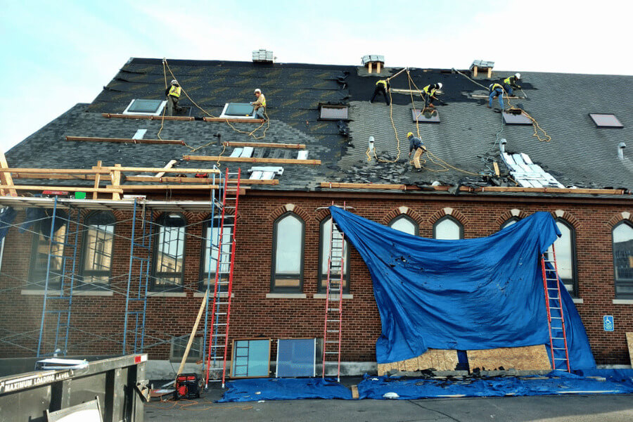 Roofing crew works across the top of a large brick building, with ladders and tarps in place during a full roof replacement project.
