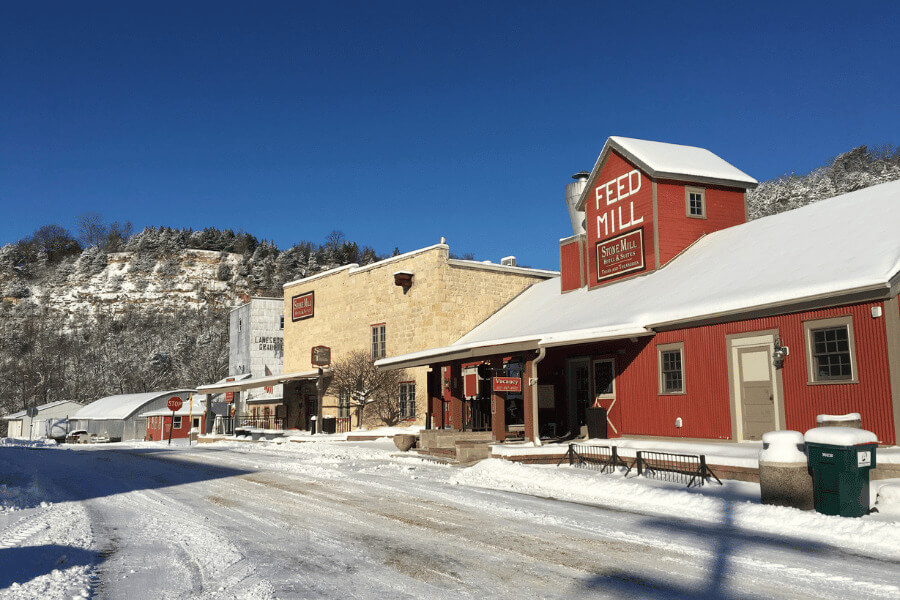 Winter street view of Stone Mill Hotel & Suites in Lanesboro, Minnesota, with the historic limestone mill and red Feed Mill building covered in fresh snow, set against a backdrop of bluffs and a clear blue sky.