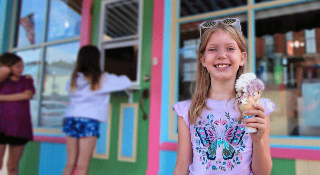 A smiling girl holding a large ice cream cone stands in front of a colorful storefront with pink, green, and blue trim. Two other children are near a walk-up service window in the background.