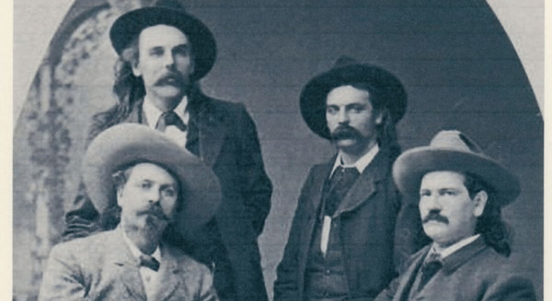 Black and white vintage photo of four men from the late 1800s, all wearing wide-brimmed hats and suits. Two are seated and two are standing behind them. The man on the left is wearing a light-colored suit and cowboy hat; the others are in darker suits. They have mustaches and long hair, styled in the fashion of the Old West. The background features an arched doorway.