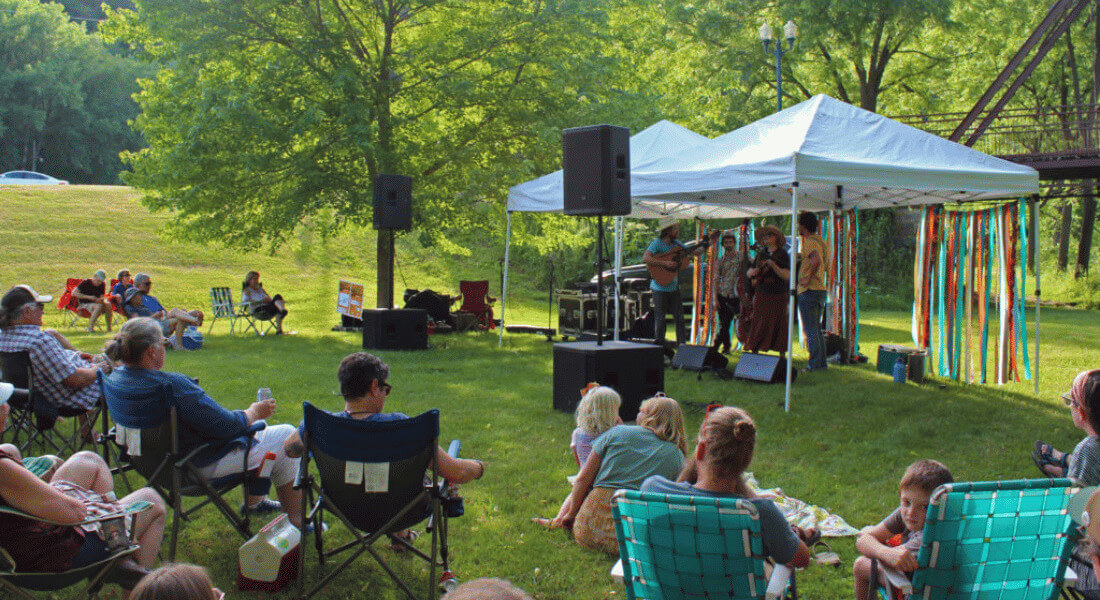 People sit on lawn chairs and blankets enjoying an outdoor summer concert in a grassy park. A small band performs under a white canopy tent decorated with colorful streamers, with trees and a bridge in the background.