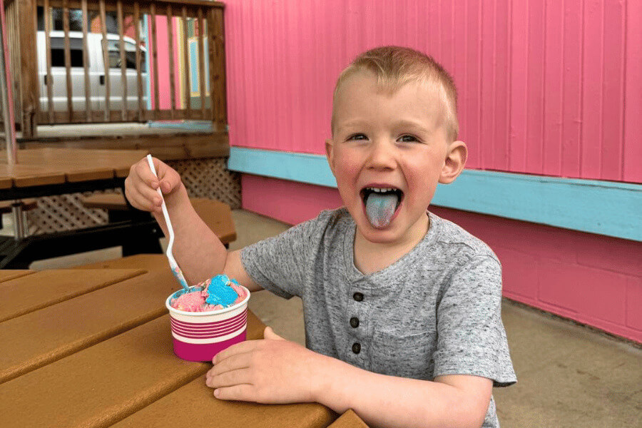 A smiling young boy with a blue tongue sits at an outdoor picnic table, holding a cup of colorful ice cream with a spoon. He’s wearing a gray shirt and is in front of a bright pink and teal wall, creating a cheerful summer vibe.