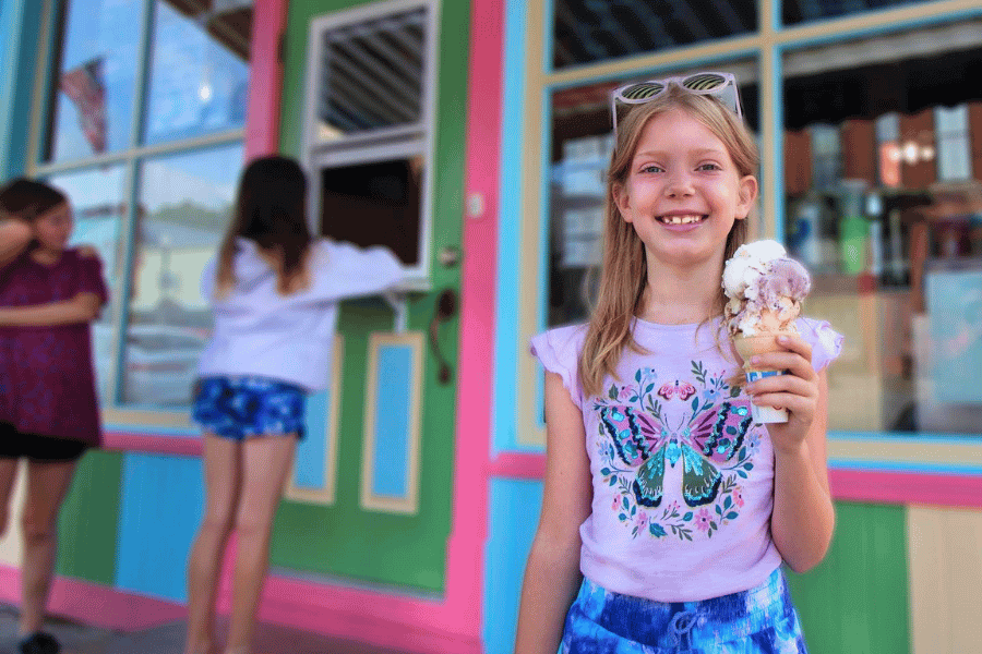 A smiling girl holding a large ice cream cone stands in front of a colorful storefront with pink, green, and blue trim. Two other children are near a walk-up service window in the background.