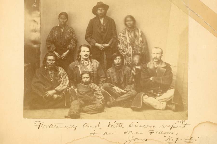 Sepia-toned 1879 photograph showing Buffalo Bill Cody and Dr. Frank “White Beaver” Powell seated with a group of Native American men, women, and a child; handwritten inscription along the bottom includes the date and a note signed "Noⁿp Ska."