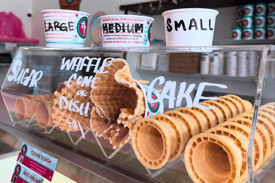 A close-up of an ice cream counter display at Clara’s Eatery showing a variety of cone options including sugar, waffle, and cake cones, with labeled sample cups for small, medium, and large sizes.
