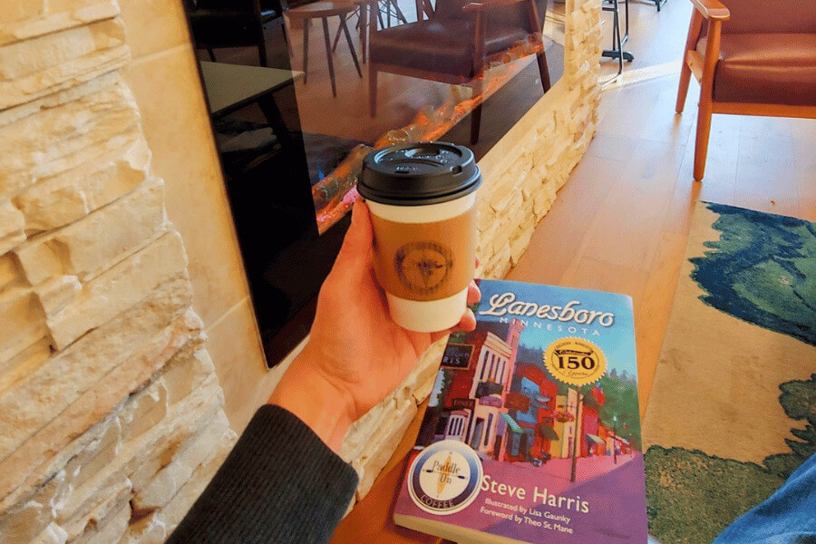 A person holding a coffee next to a fireplace with the book "Lanesboro, MN" visible