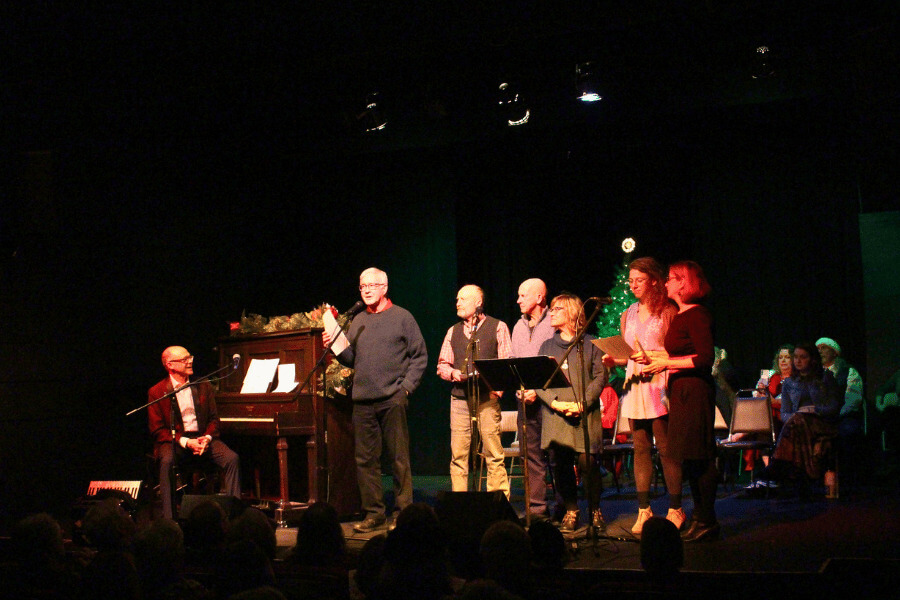 A group of people stands on stage singing during a holiday sing-along at the St. Mane Theatre. Dan Chouinard plays the piano beside them, with festive decorations and a lit Christmas tree in the background. Audience members watch from the darkened theater.