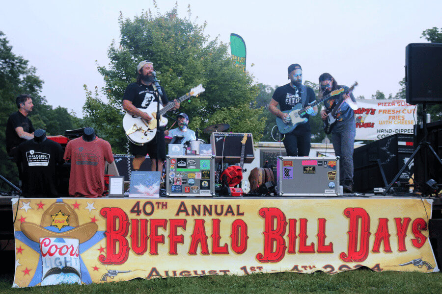 A band performs on an outdoor stage at the 40th Annual Buffalo Bill Days festival, with musicians playing guitars and singing into microphones. The stage is decorated with a large banner displaying the event name and date, and merchandise is set up in front of the performers. Trees and food stands are visible in the background.
