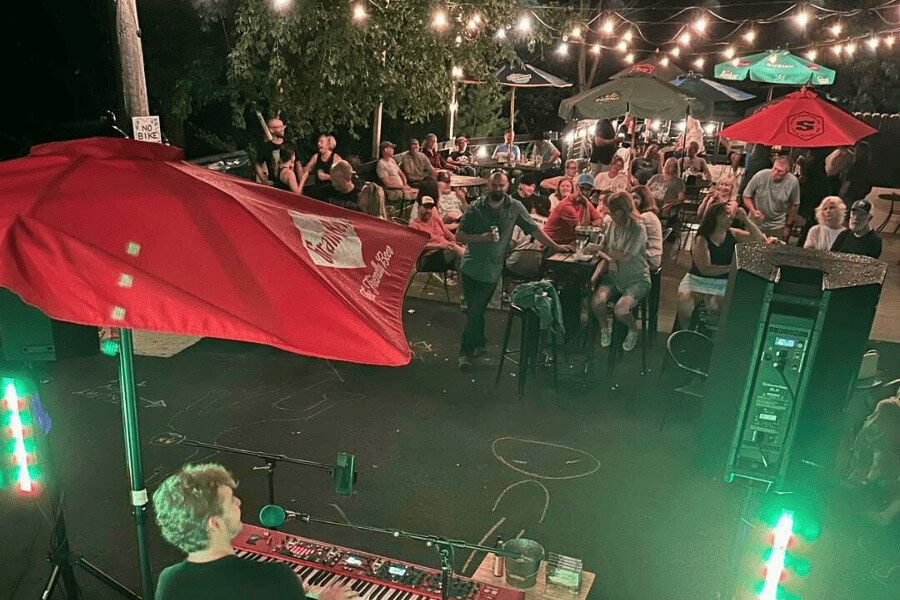 A musician plays keyboard and sings into a microphone during an outdoor nighttime performance at a lively pub patio. The audience, seated at tables under large umbrellas and string lights, enjoys the music and atmosphere.