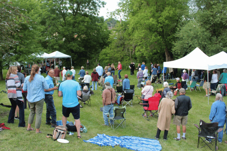 A crowd gathers on a grassy lawn to enjoy a live outdoor concert. People stand, sit in lawn chairs, or relax on blankets while a band performs under a white tent stage surrounded by lush green trees.
