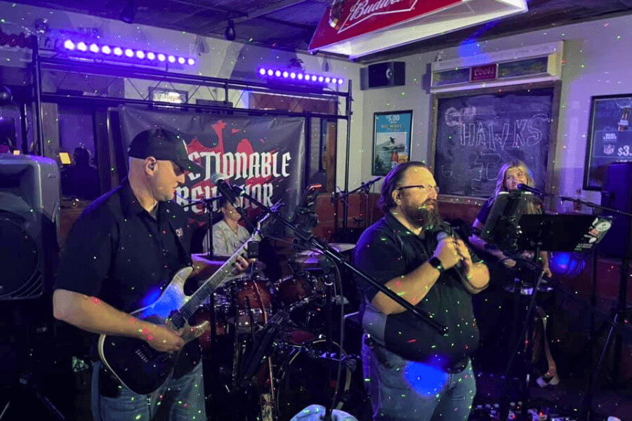 A band performs live at an indoor venue with colorful stage lighting. The guitarist, drummer, lead vocalist, and keyboardist are mid-performance in front of a banner reading “Questionable Behavior.” The audience is not visible, but the cozy bar atmosphere is evident.