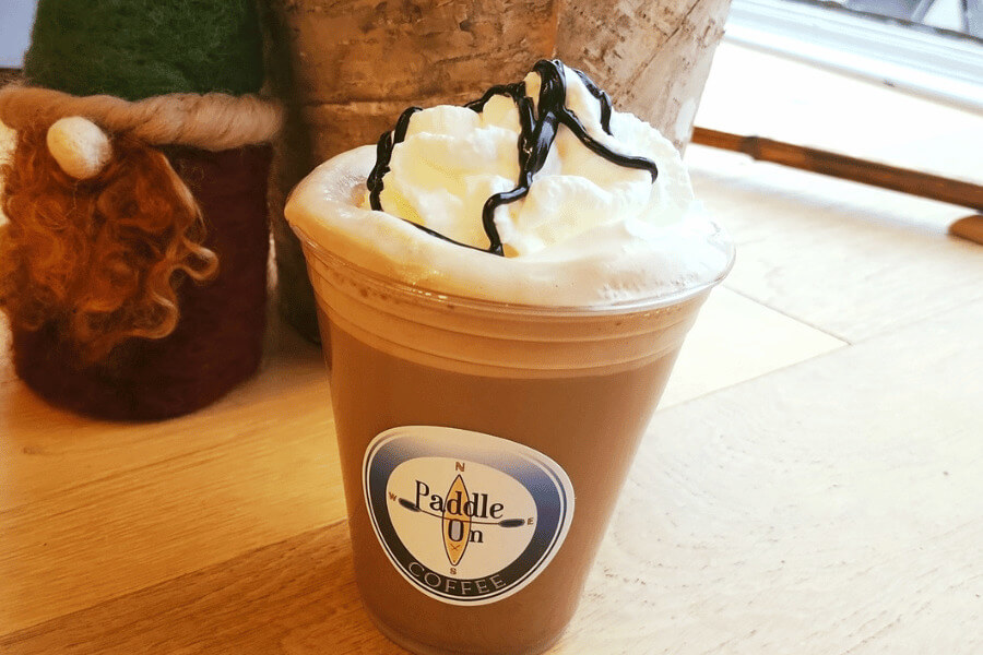 A sweet iced mocha topped with whipped cream and chocolate drizzle sits on a wooden table at Paddle On Coffee in Lanesboro. A cozy felt gnome and rustic birch decor add a charming touch in the background.