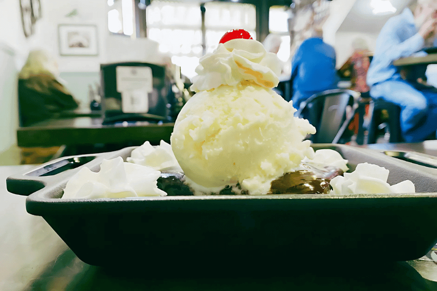 A decadent brownie sundae topped with a generous scoop of vanilla ice cream, whipped cream, chocolate sauce, and a cherry is served in a black dish at Pedal Pushers Café, with diners in the background.