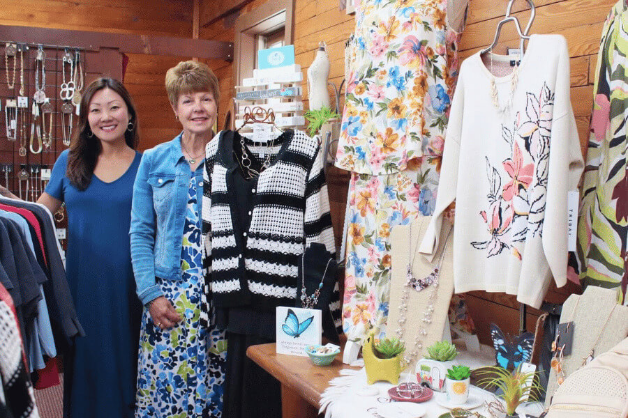 Annie and Therese standing inside Stone Mill Clothing & Gifts in Lanesboro, Minnesota, surrounded by women’s apparel, jewelry, and accessories, including floral dresses, knit sweaters, and display tables with gift items.