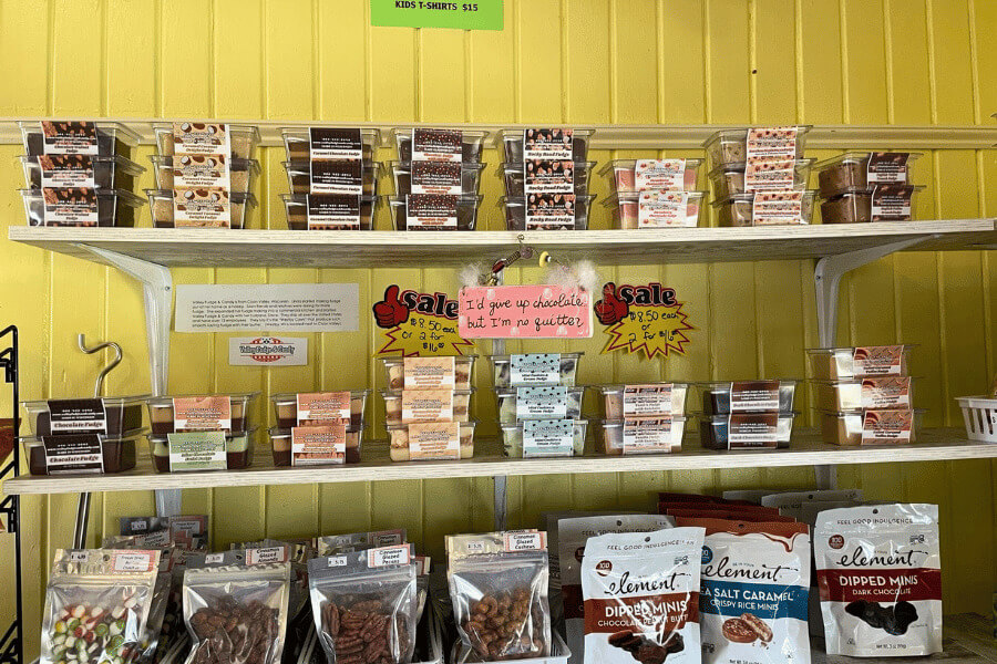 A colorful display of assorted fudge and candy packs is arranged neatly on white shelves against a bright yellow wall at the Whalan Depot.