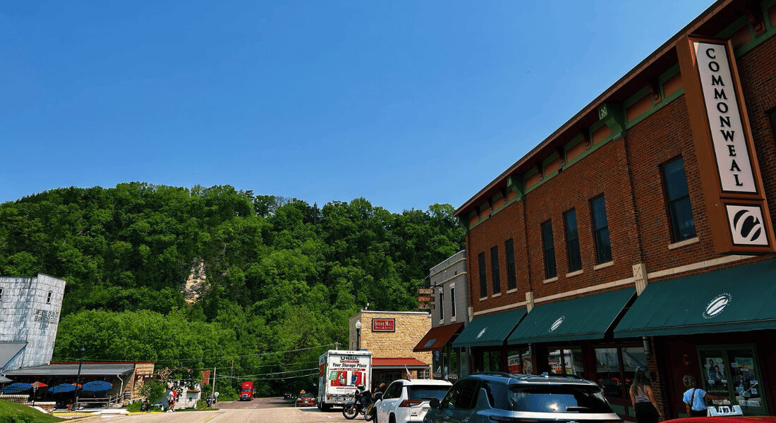View of downtown Lanesboro, Minnesota, with the Commonweal Theatre building in the foreground and tree-covered bluffs in the background on a clear summer day.