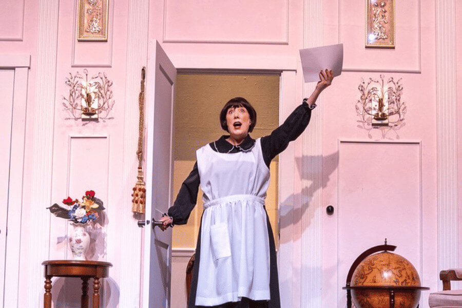 Actor in a maid costume dramatically enters a brightly lit stage set resembling an elegant living room, holding a piece of paper and striking a theatrical pose.