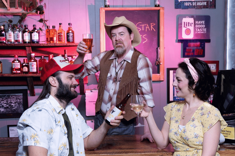 Three people toast drinks in a small-town bar, with liquor bottles and neon signs in the background.