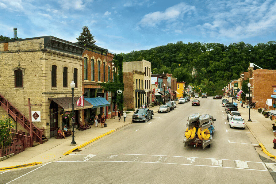 Downtown Lanesboro, MN with historic buildings, American flags, and a truck hauling kayaks and tubes—set against lush green bluff country under a blue sky. Photo by Explore Minnesota / Ryan Taylor.