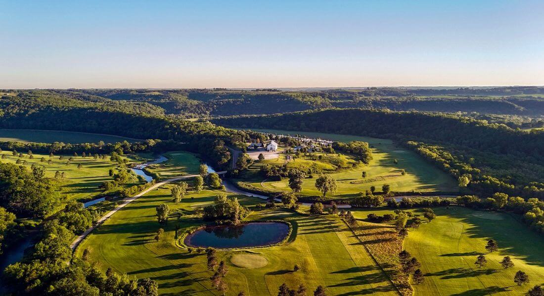 Aerial view of the Old Barn Resort and Rivers Bend Golf Course near Lanesboro, Minnesota, surrounded by rolling green hills, tree-lined fairways, a winding river, and a small pond under clear blue skies.