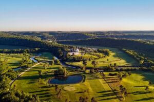 Aerial view of the Old Barn Resort and Rivers Bend Golf Course near Lanesboro, Minnesota, surrounded by rolling green hills, tree-lined fairways, a winding river, and a small pond under clear blue skies.