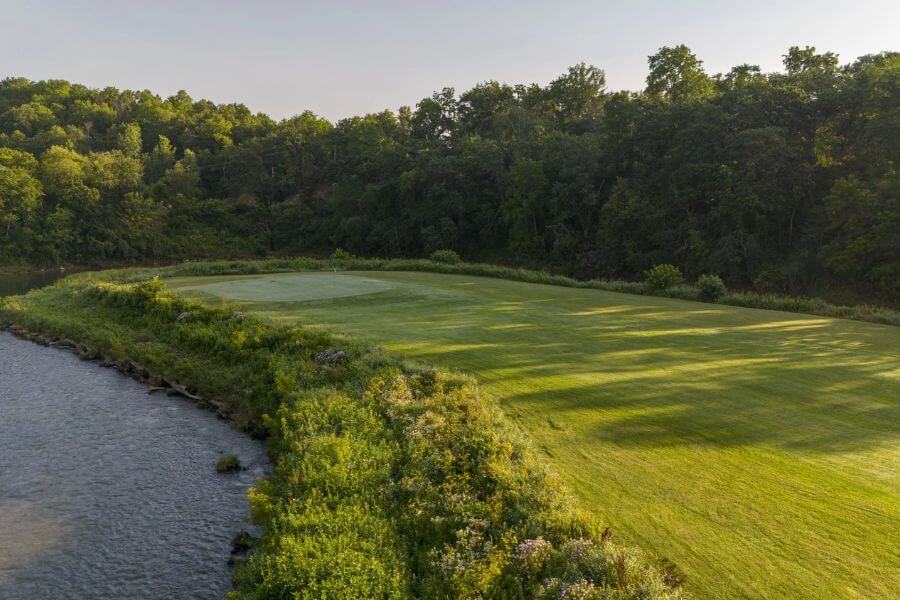 A golf green at Rivers Bend Golf Course sits right along the edge of the Root River, surrounded by lush trees and morning sunlight.