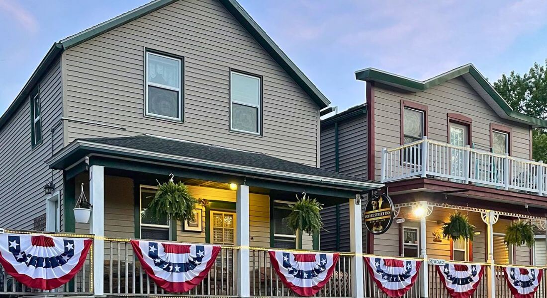 Exterior of Coffee Street Inn in Lanesboro, Minnesota, decorated with red, white, and blue bunting and hanging ferns, with the inn’s sign visible on the porch at dusk.