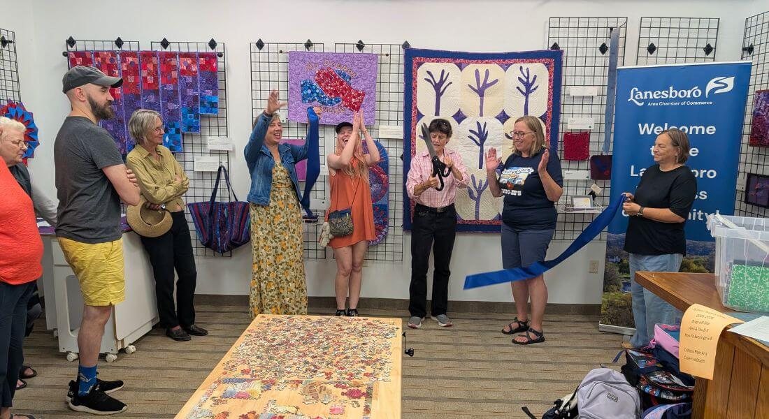 Community members and leaders gather inside Driftless Fiber Arts Collective for a ribbon cutting ceremony, surrounded by colorful fiber art and quilts on display.