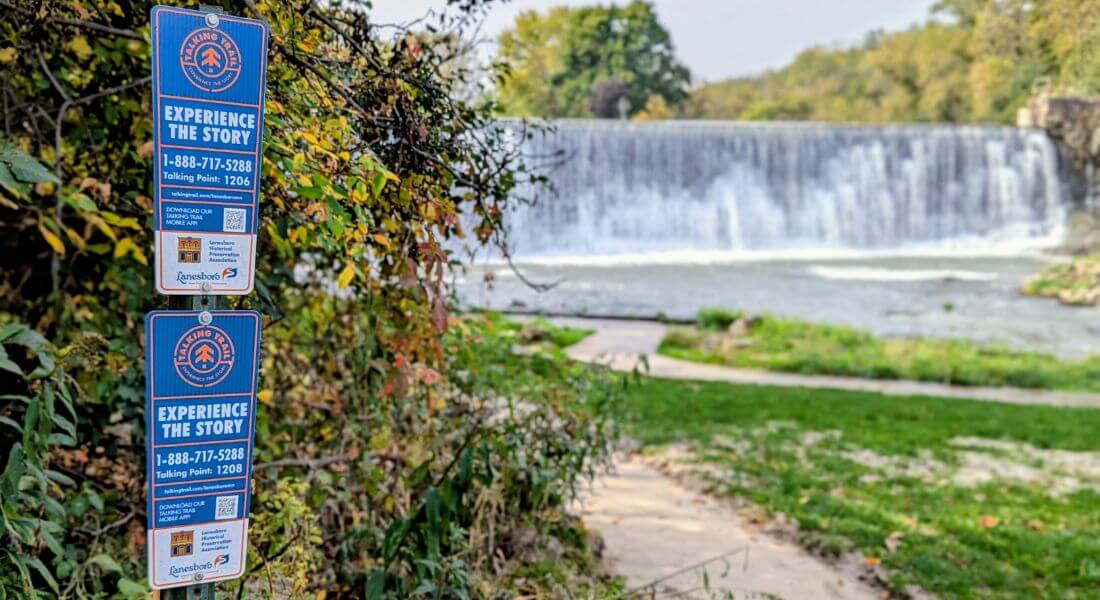Talking Trail signs posted near the Lanesboro Dam, with the waterfall flowing in the background along the Root River.