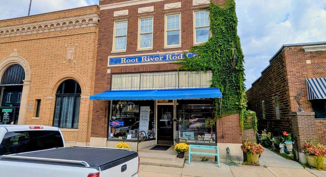 Front entrance of Root River Rod Co. in downtown Lanesboro, Minnesota, with a bright blue awning, ivy climbing the brick wall, and yellow mums by the doorway.