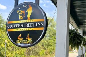 Round black-and-gold Coffee Street Inn sign in Lanesboro, Minnesota, featuring graphics of a fisherman and cyclists, hanging outside the inn against a backdrop of trees and blue sky.