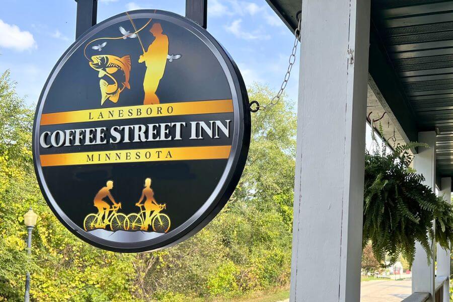 Round black-and-gold Coffee Street Inn sign in Lanesboro, Minnesota, featuring graphics of a fisherman and cyclists, hanging outside the inn against a backdrop of trees and blue sky.