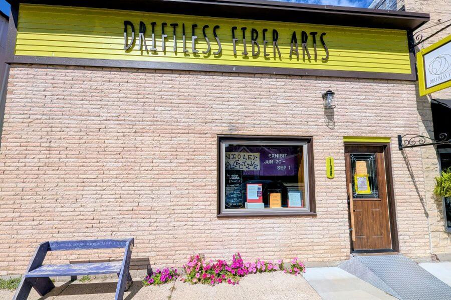 Exterior of the Driftless Fiber Arts Collective building in downtown Lanesboro, with a bright yellow sign above the entrance and window posters promoting the Violet Collective exhibition.