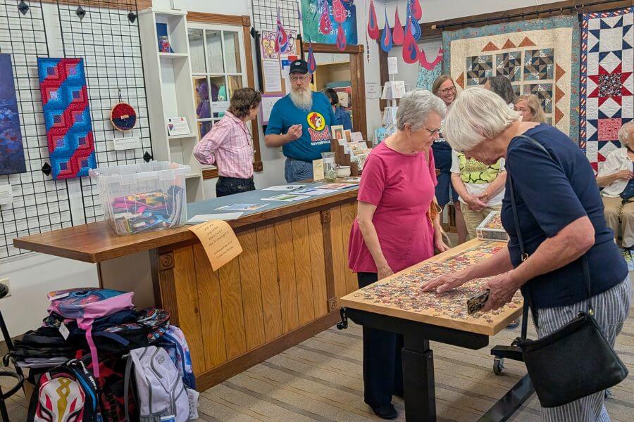 Guests at the Driftless Fiber Arts Collective engage with a large community puzzle table while others browse fiber art and handmade items on display.