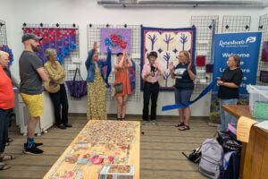 Community members and leaders gather inside Driftless Fiber Arts Collective for a ribbon cutting ceremony, surrounded by colorful fiber art and quilts on display.