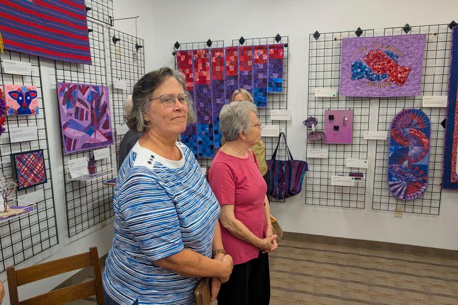 Visitors view colorful fiber art pieces made from red, blue, and violet materials during the Violet Collective exhibition at Driftless Fiber Arts Collective.
