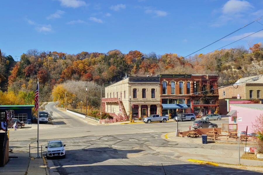 View of downtown Lanesboro, MN on a sunny autumn day. Historic brick buildings line the street with colorful fall foliage covering the bluffs in the background. Cars are parked along the street, and the American flag waves near the corner.