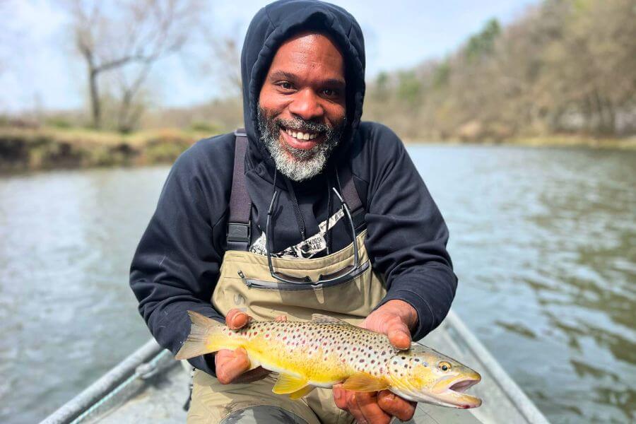 Smiling angler in waders holding a freshly caught brown trout while standing in a boat on the Root River near Lanesboro, Minnesota.