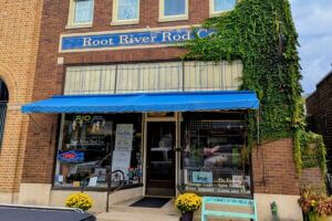Front entrance of Root River Rod Co. in downtown Lanesboro, Minnesota, with a bright blue awning, ivy climbing the brick wall, and yellow mums by the doorway.
