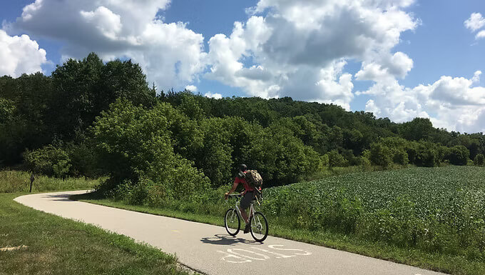 Cyclist riding on the Root River State Trail near Lanesboro, Minnesota, surrounded by green fields, trees, and summer skies.