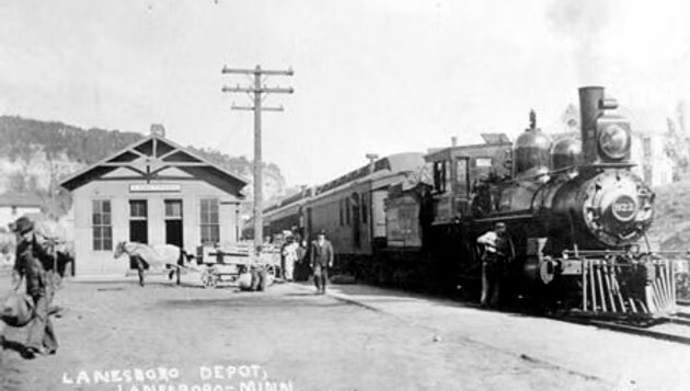 Historic photo of a steam locomotive at the Lanesboro Depot in Minnesota during the railroad era, with passengers, workers, and horse-drawn wagons nearby.