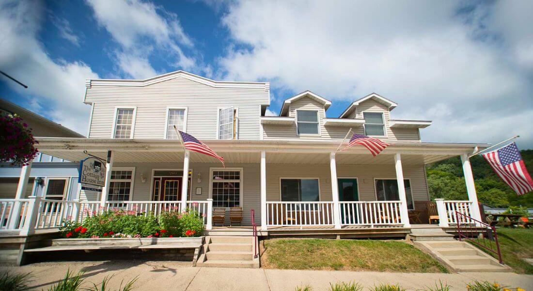 Front exterior of The Cottage House Inn in downtown Lanesboro, Minnesota, featuring a wide front porch with rocking chairs, American flags, and blooming flowers under a bright summer sky.