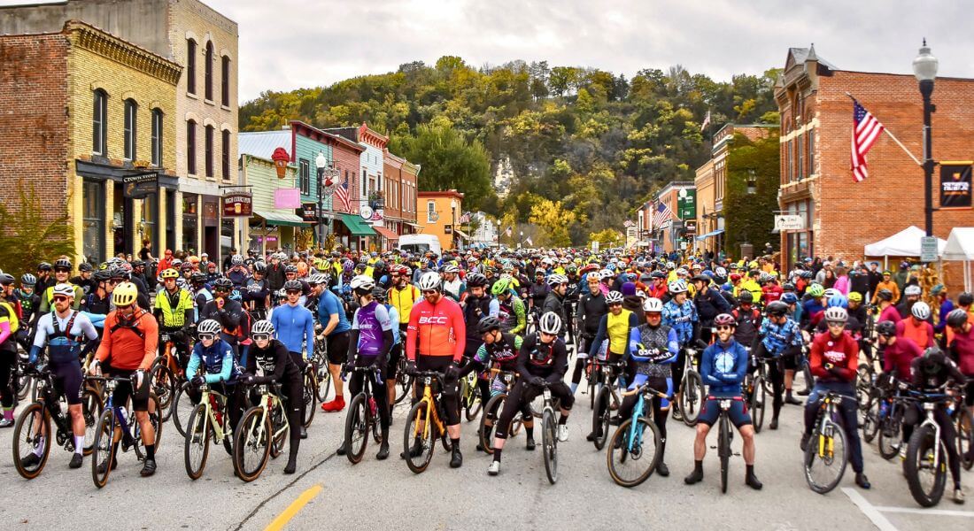 Hundreds of cyclists line up on Lanesboro’s historic downtown street for the start of the Filthy 50 Gravel Bike Race, surrounded by colorful storefronts and autumn bluff scenery.