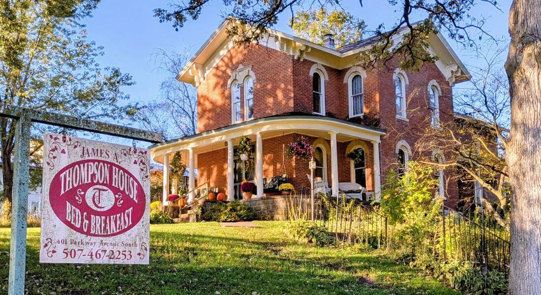 The James A. Thompson House Bed & Breakfast in Lanesboro, Minnesota—a beautifully restored 1870s Italianate brick mansion with white columns, arched windows, and a welcoming front porch decorated with pumpkins and fall flowers. A vintage-style sign in the foreground reads “James A. Thompson House Bed & Breakfast.”