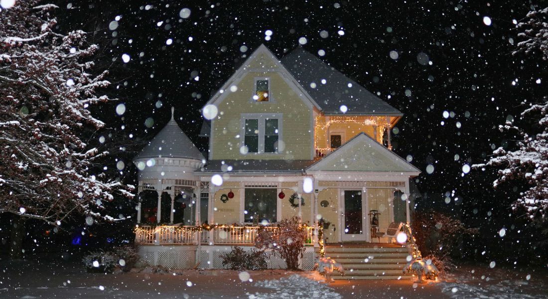 A warmly lit Victorian-style inn in Lanesboro, Minnesota, glows with holiday lights as snow gently falls at night, creating a peaceful and cozy winter scene.