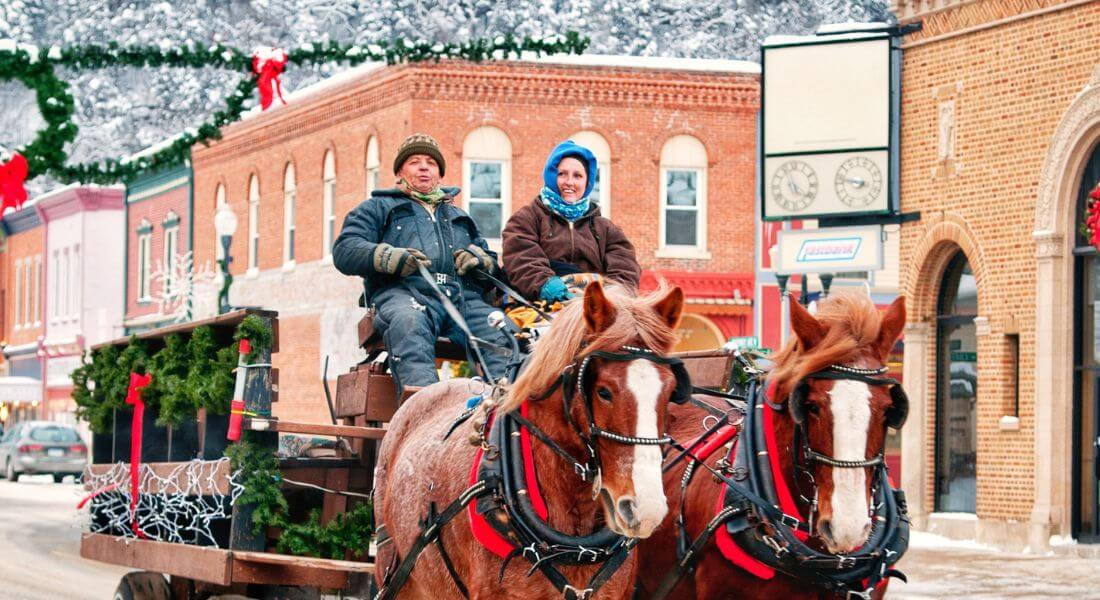 A horse-drawn carriage makes its way down Lanesboro’s snow-covered main street, driven by two warmly dressed locals. The decorated wagon, holiday wreaths, and twinkling lights create a festive small-town winter scene.
