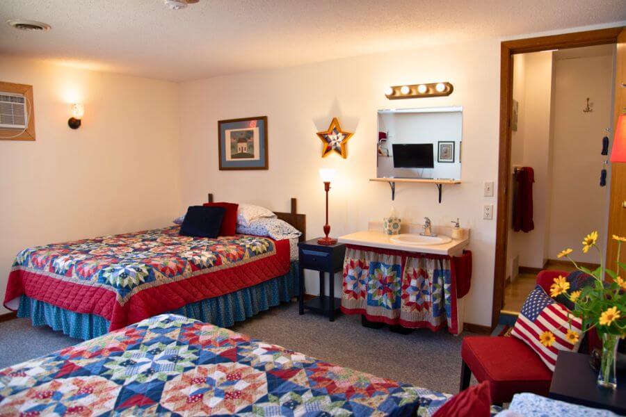 A bright, cozy guest room at A Guest Hus Motel in Lanesboro, Minnesota, featuring two beds with colorful patchwork quilts, a small vanity sink, patriotic-themed décor, and warm lighting that creates a welcoming, homey atmosphere.
