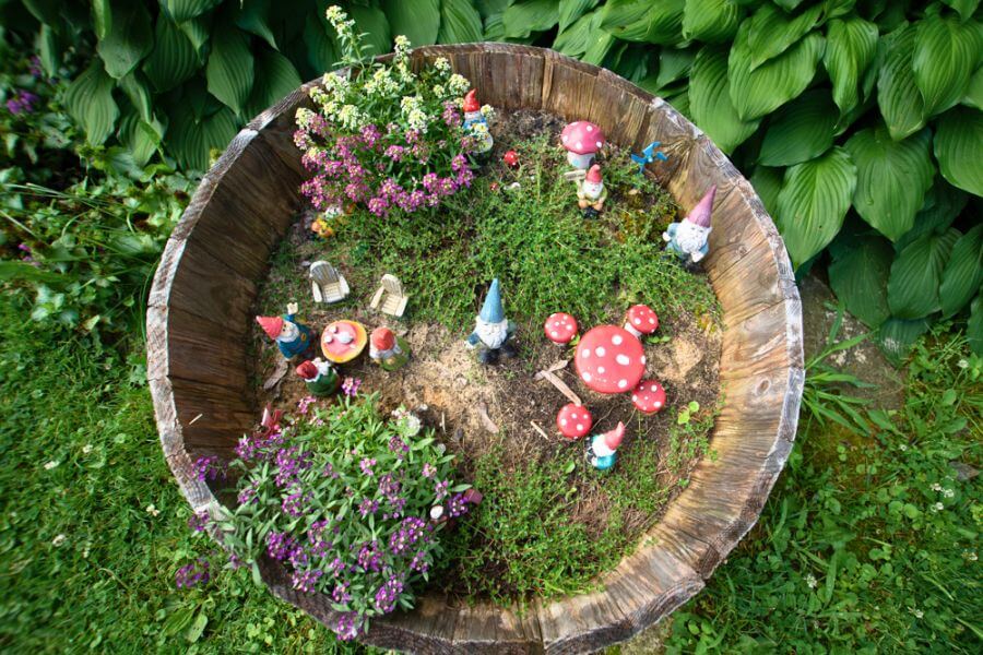 A whimsical garden display inside a wooden barrel planter featuring colorful gnome figurines, tiny mushroom decorations, and blooming flowers surrounded by lush green foliage at A Guest Hus Motel in Lanesboro, Minnesota.